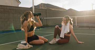 Two lasies enjoying a break from playing pickle ball sitting on the court and drinking a sunwarrior mix.