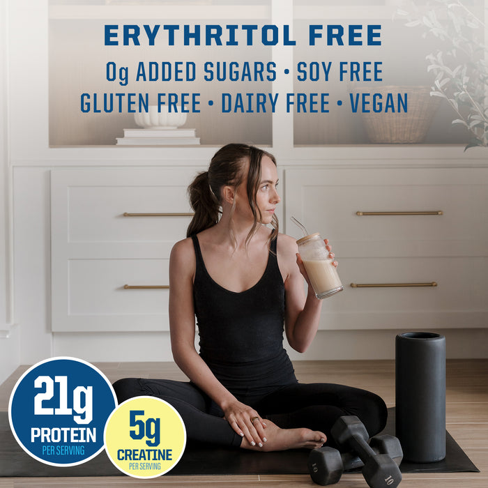 Woman in workout clothes sitting on a yoga mat holding a protein shake with weights and foam roller nearby