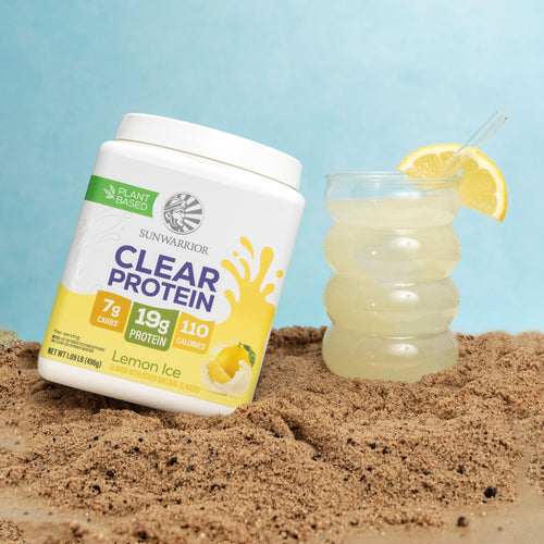 Container of lemon-flavored plant-based protein powder next to a glass of lemonade on sand.