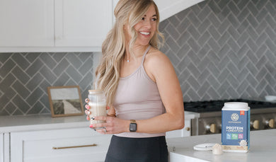 A woman in a kitchen holding a protein shake, with a blue tub of Sunwarrior Protein Creatine Powder on the counter next to her.